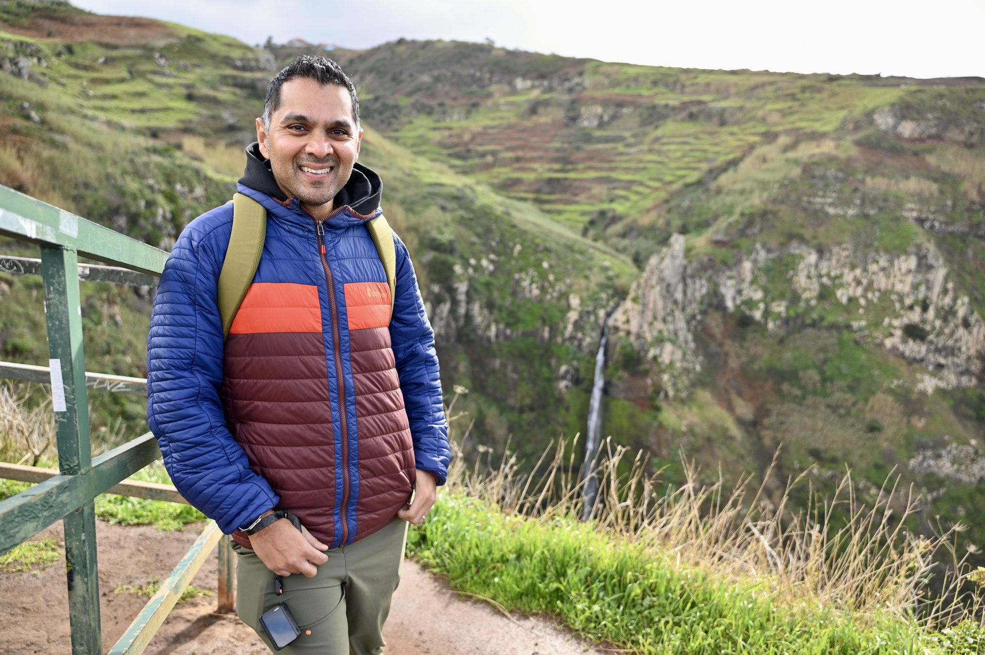 His dad smiling at the railing, with a thin waterfall dropping down a green terraced gorge behind him.