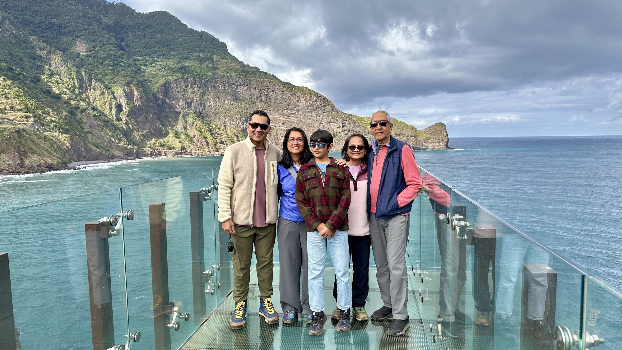 Five of us standing on the glass platform, with the sea and green mountains behind.