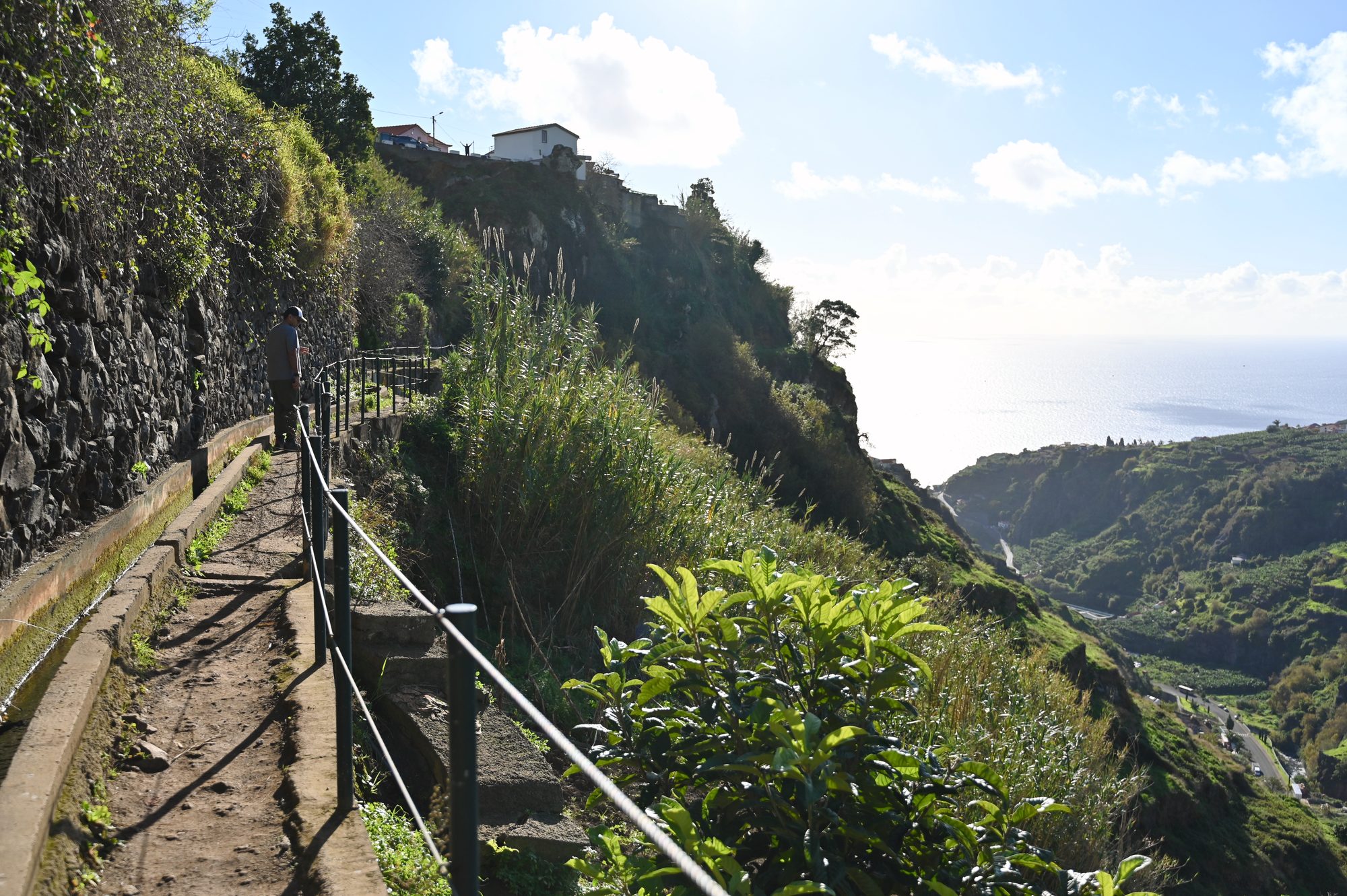 A narrow path beside the levada channel, with a railing, lush plants and the sea shining in the distance.