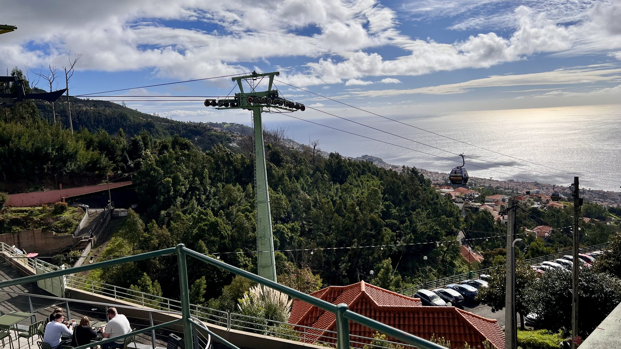 A cable car gondola heading down over green hills towards the sea, seen past a cable car pylon.