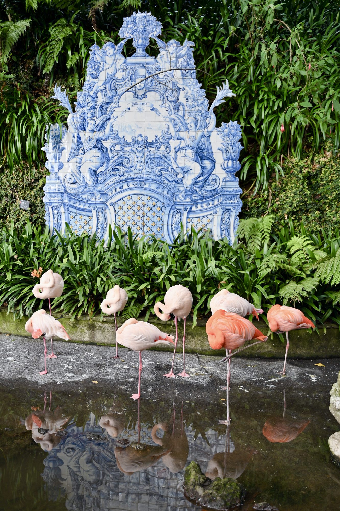A large ornate panel of blue and white azulejo tiles among ferns, with pink flamingos standing in the pond in front.
