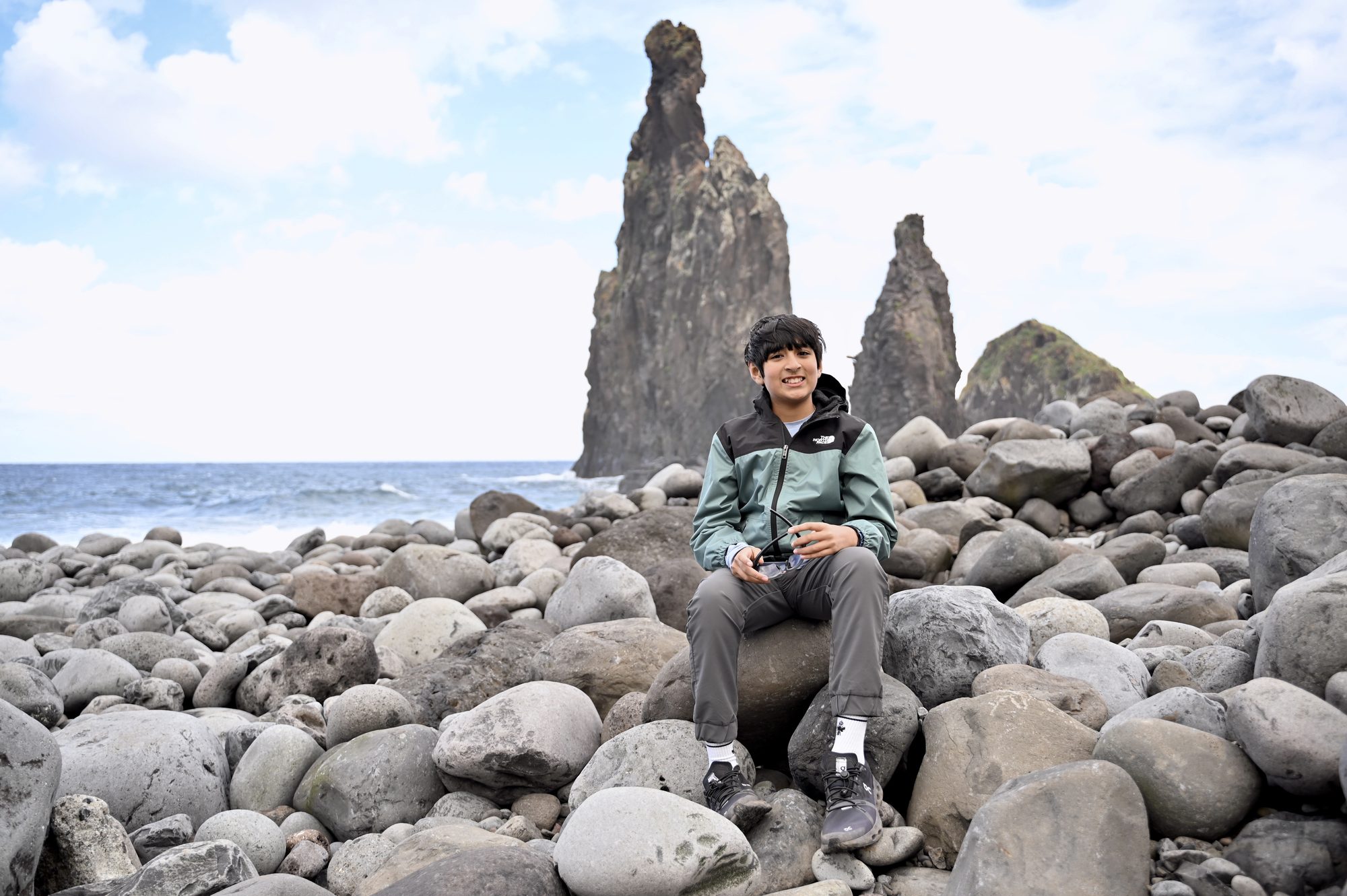 Our son sitting on smooth grey boulders, with tall, jagged sea stacks rising out of the ocean behind him.