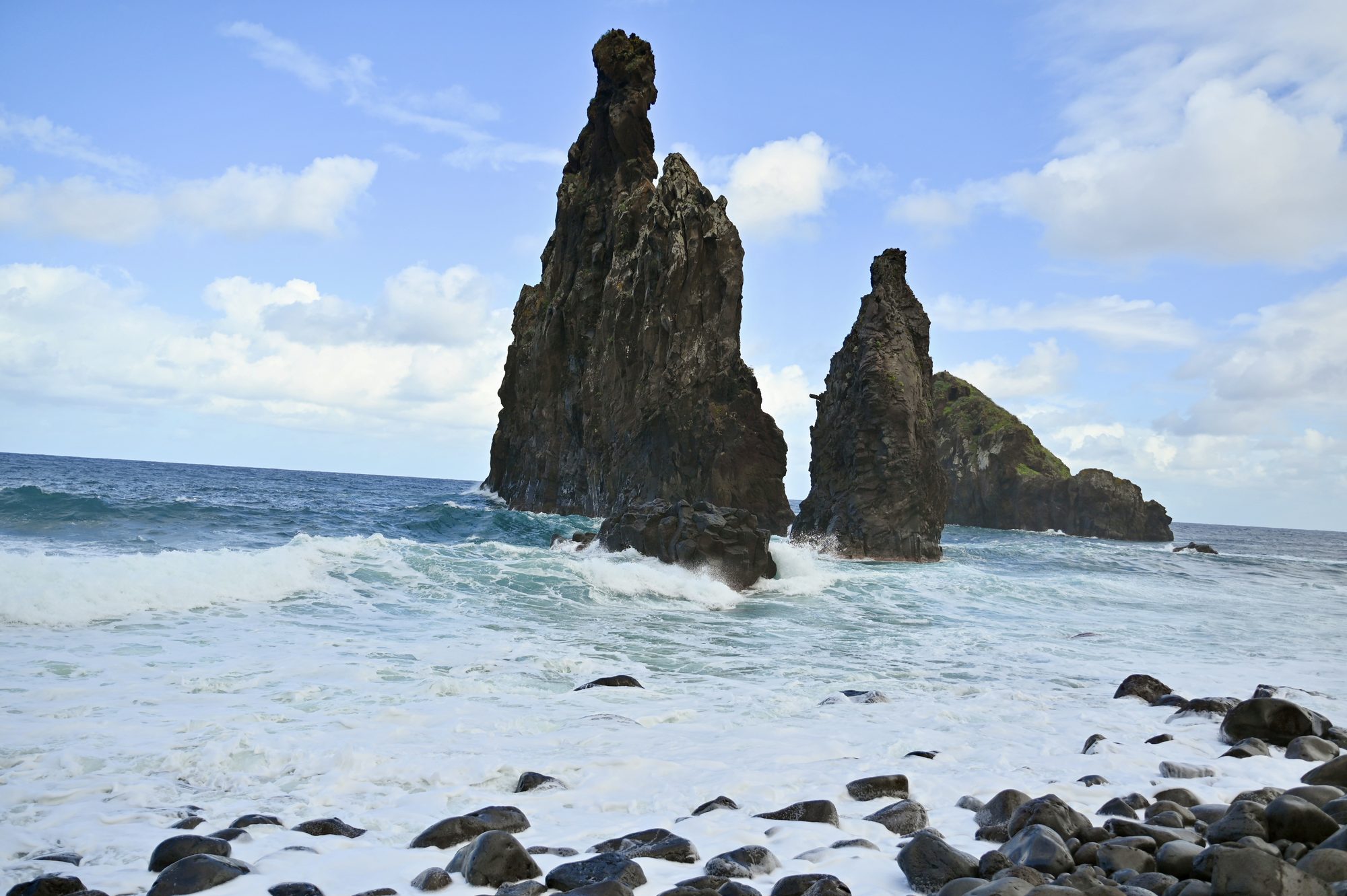 Two dark sea stacks with waves crashing around their bases, under a blue sky with clouds.
