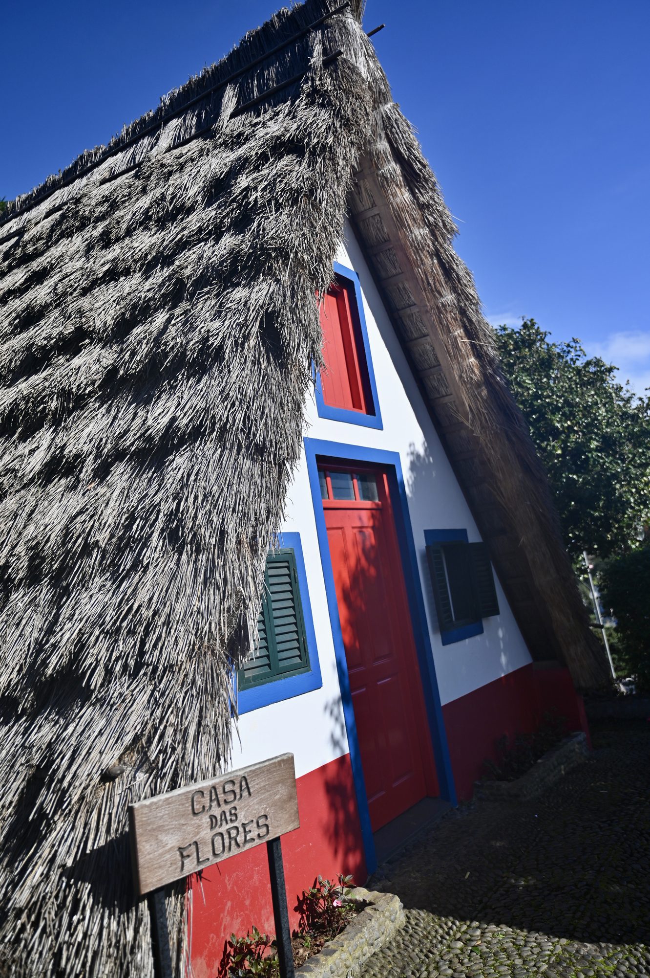 A traditional triangular Santana house with a thatched roof, red door and blue trim, and a sign reading “Casa das Flores”.