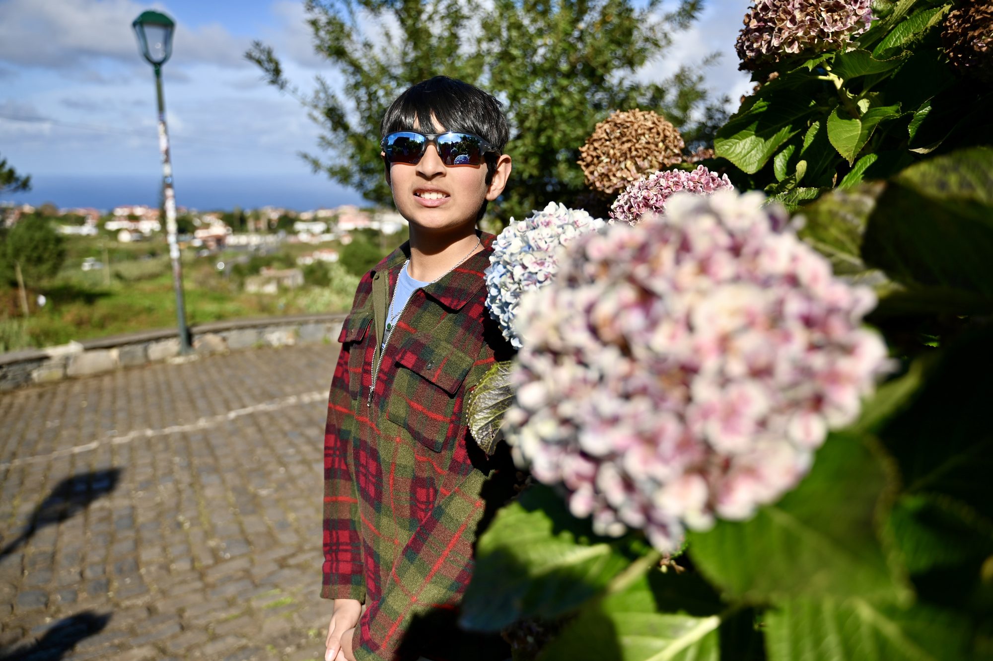Our son in sunglasses beside a big pink hydrangea, with the sea in the distance.