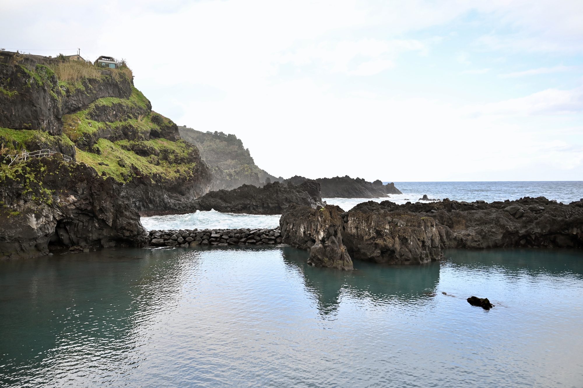 A calm, clear pool enclosed by dark volcanic rock, with the open sea beyond.
