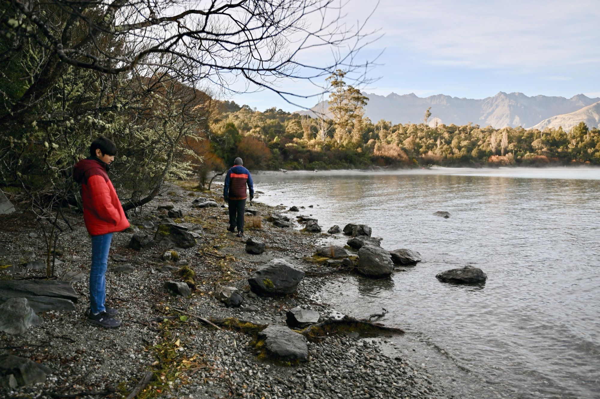 Our son and his dad walking along a rocky lakeshore under bare trees, with mountains across the water.