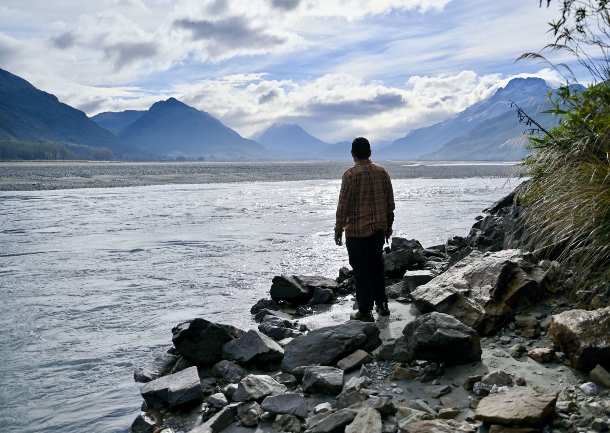 His dad standing on rocks at the edge of a wide braided river, looking towards a valley of mountains under clouds.
