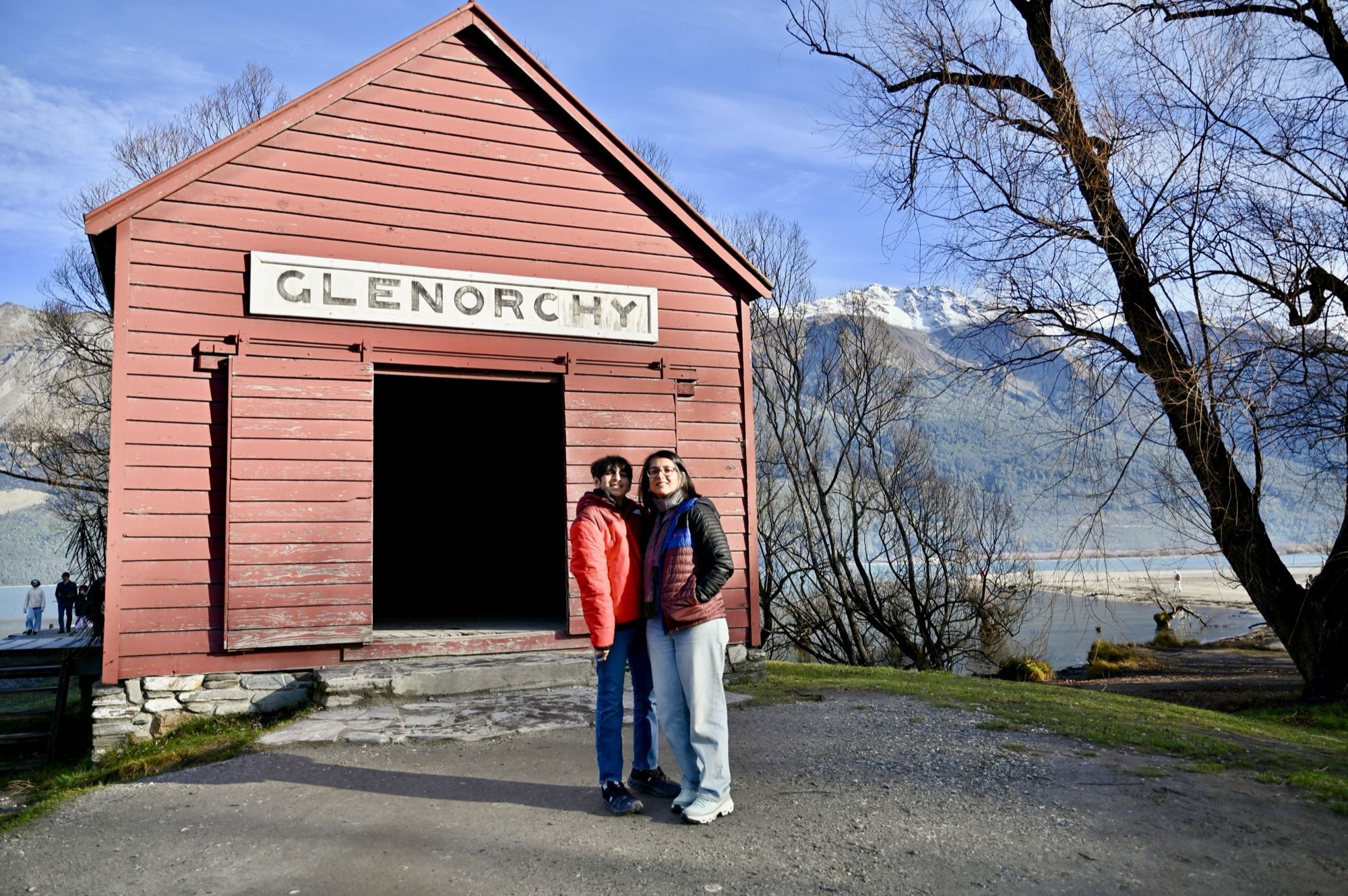 Our son and I in front of a small red wooden shed with a “Glenorchy” sign, with bare trees and mountains behind.