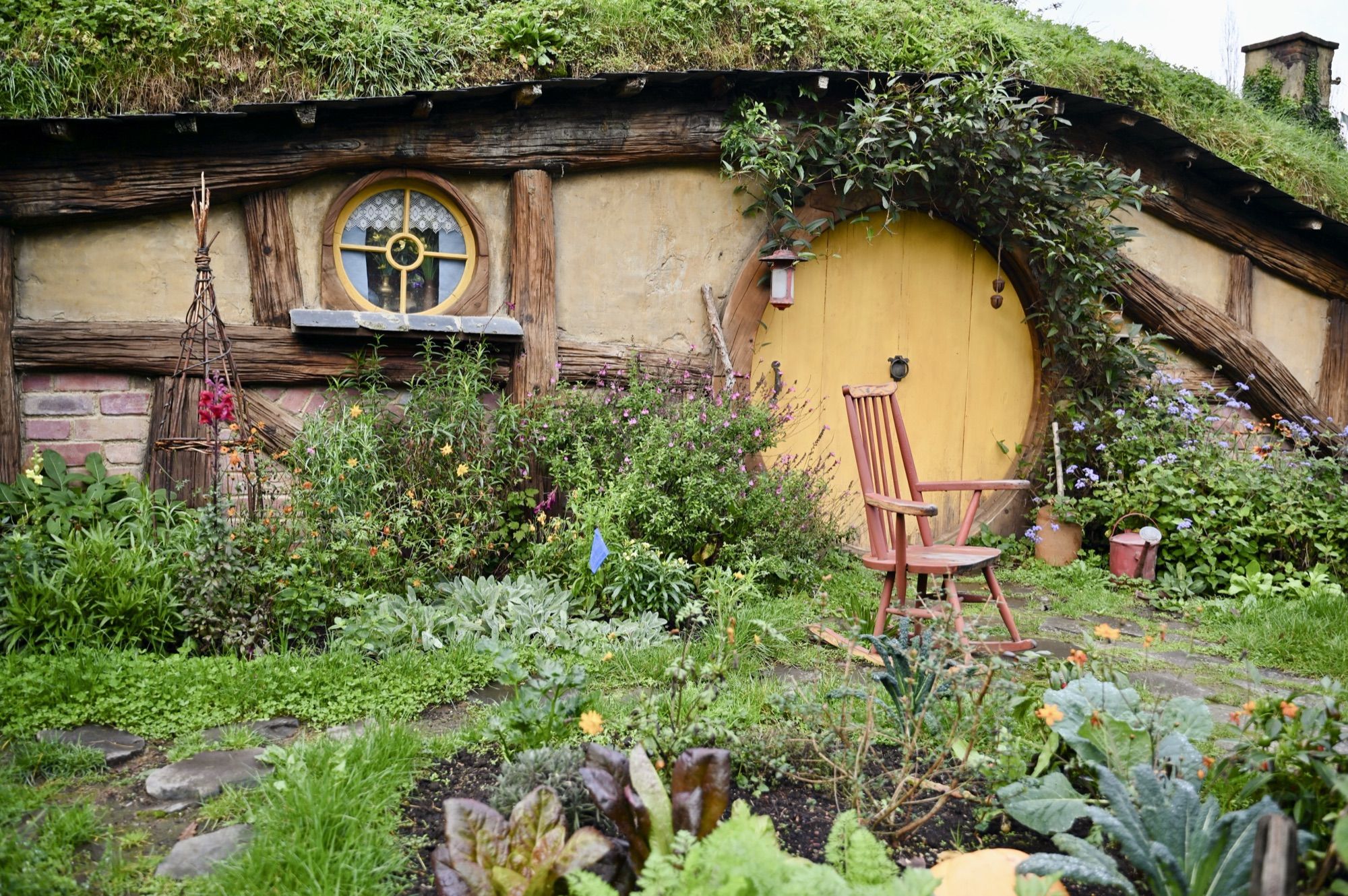 A hobbit hole with a round yellow door and round window set into a grassy hillside, with a vegetable garden and a wooden chair in front.