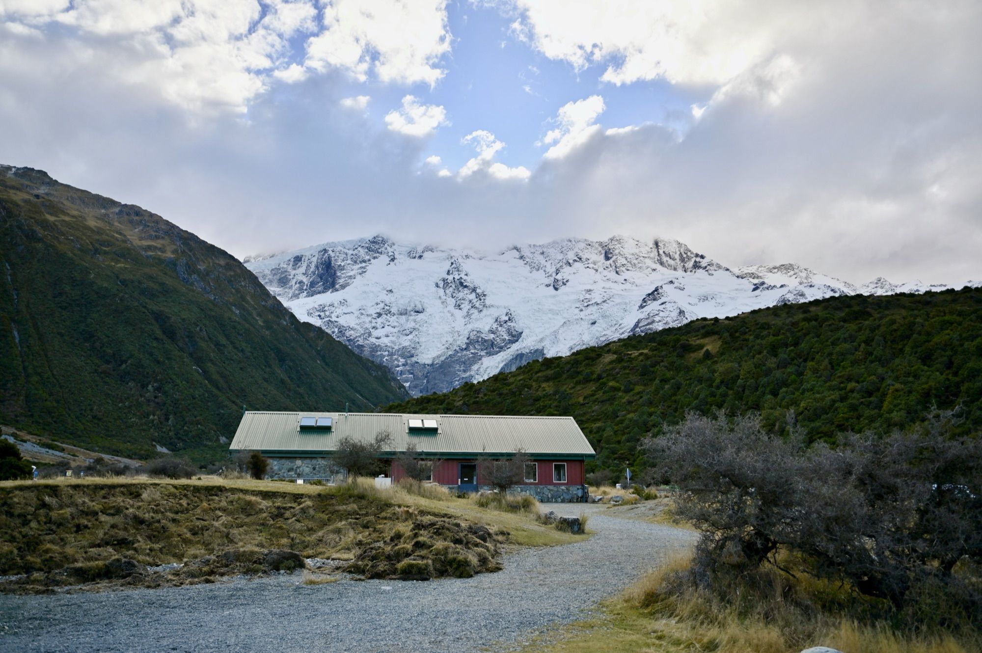 A low building at the end of a gravel road, with snow-covered mountains above the valley.