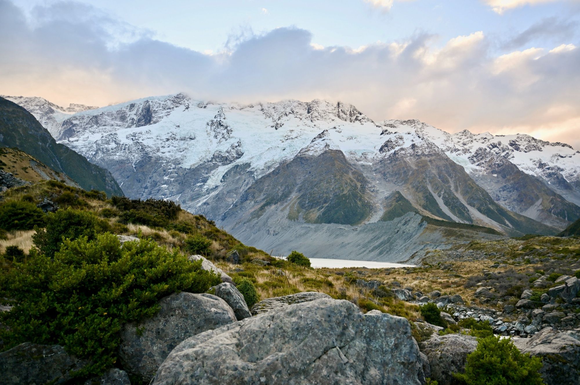 A valley of boulders and green shrubs leading to a glacier lake below a wall of snowy mountains.