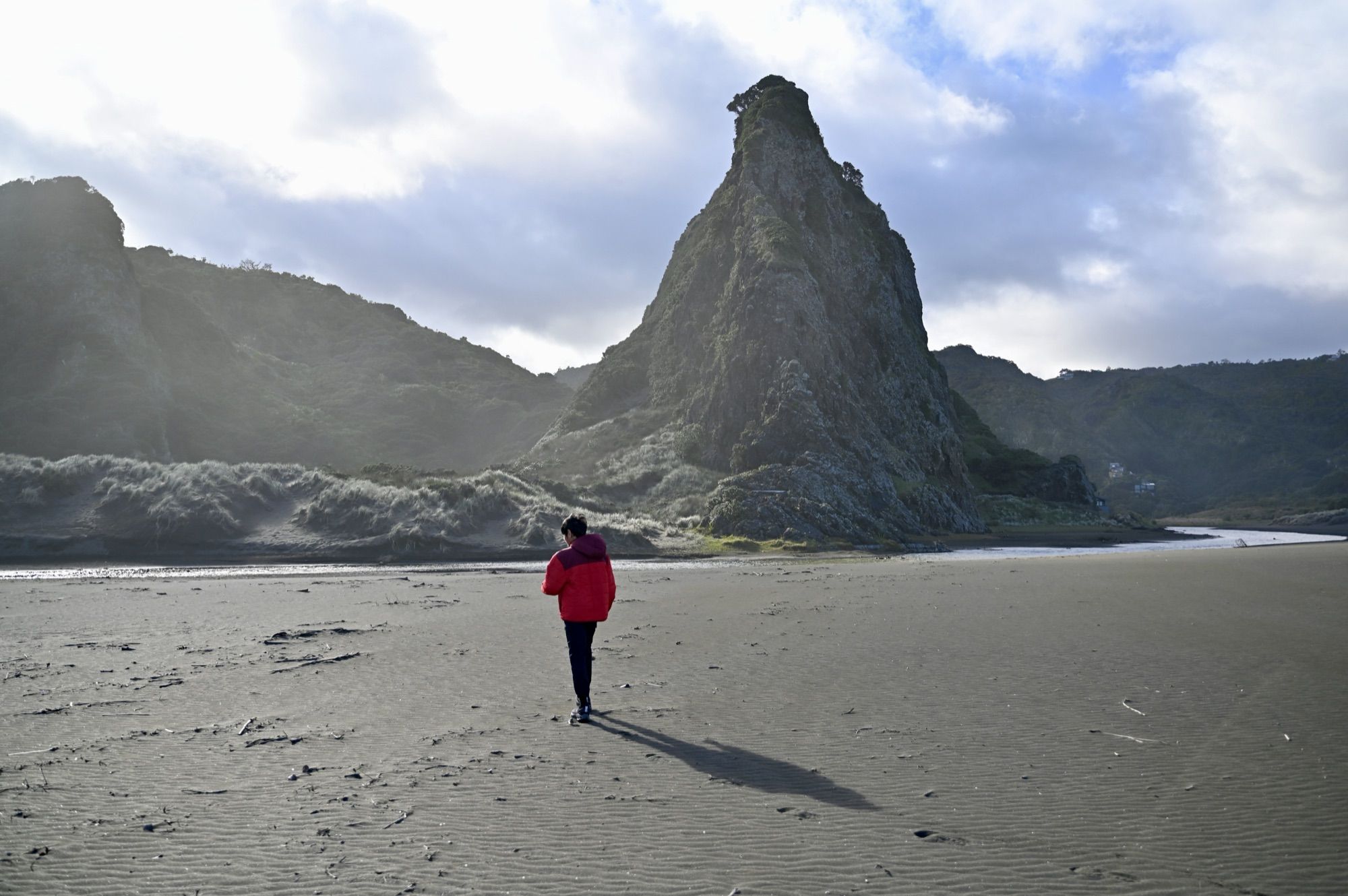 Our son in a red jacket walking across wide, dark sand towards a towering rock outcrop at Karekare Beach.