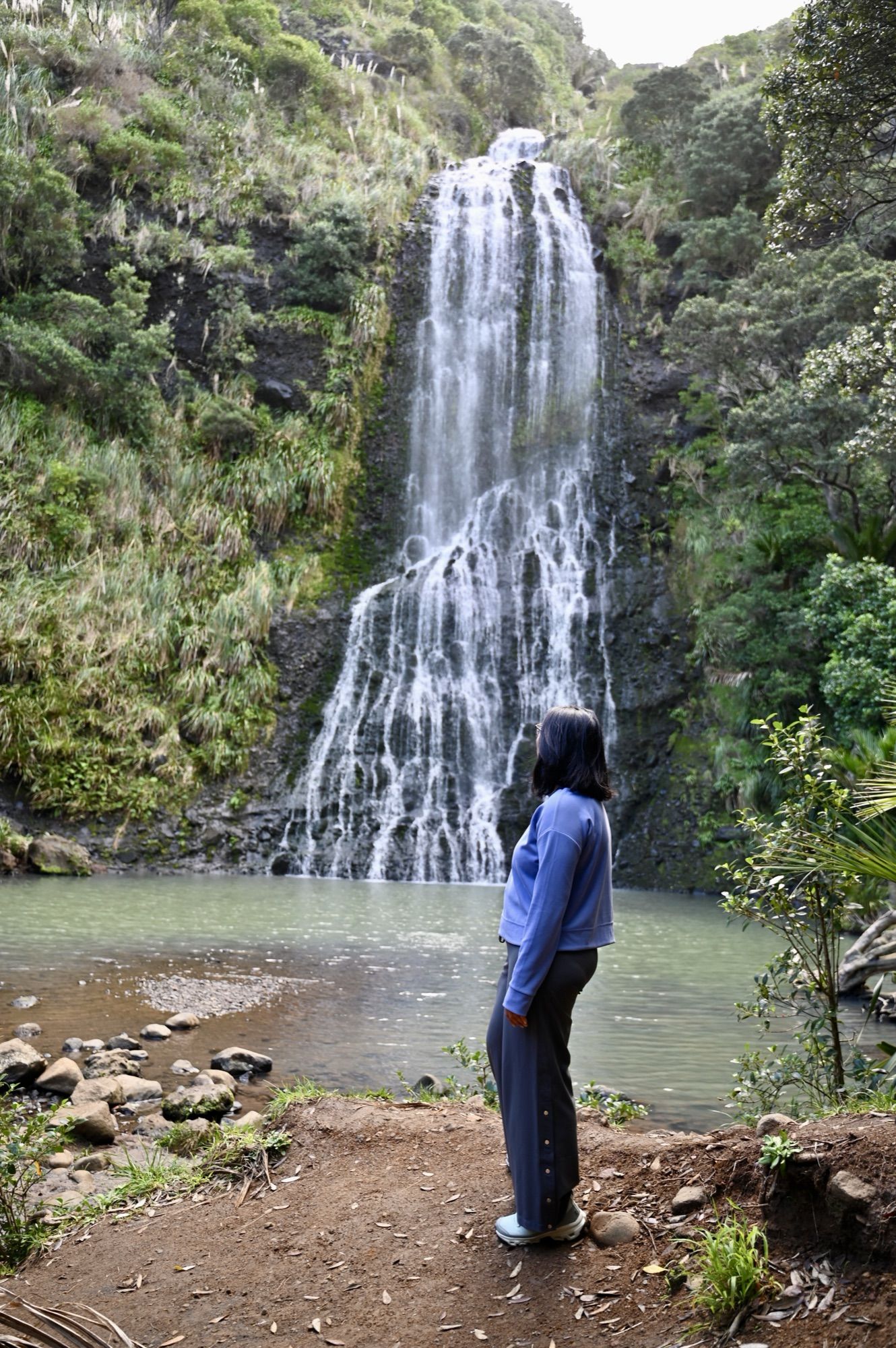 Me standing beside the pool at the base of Karekare Falls, which pours down a cliff covered in green bush.