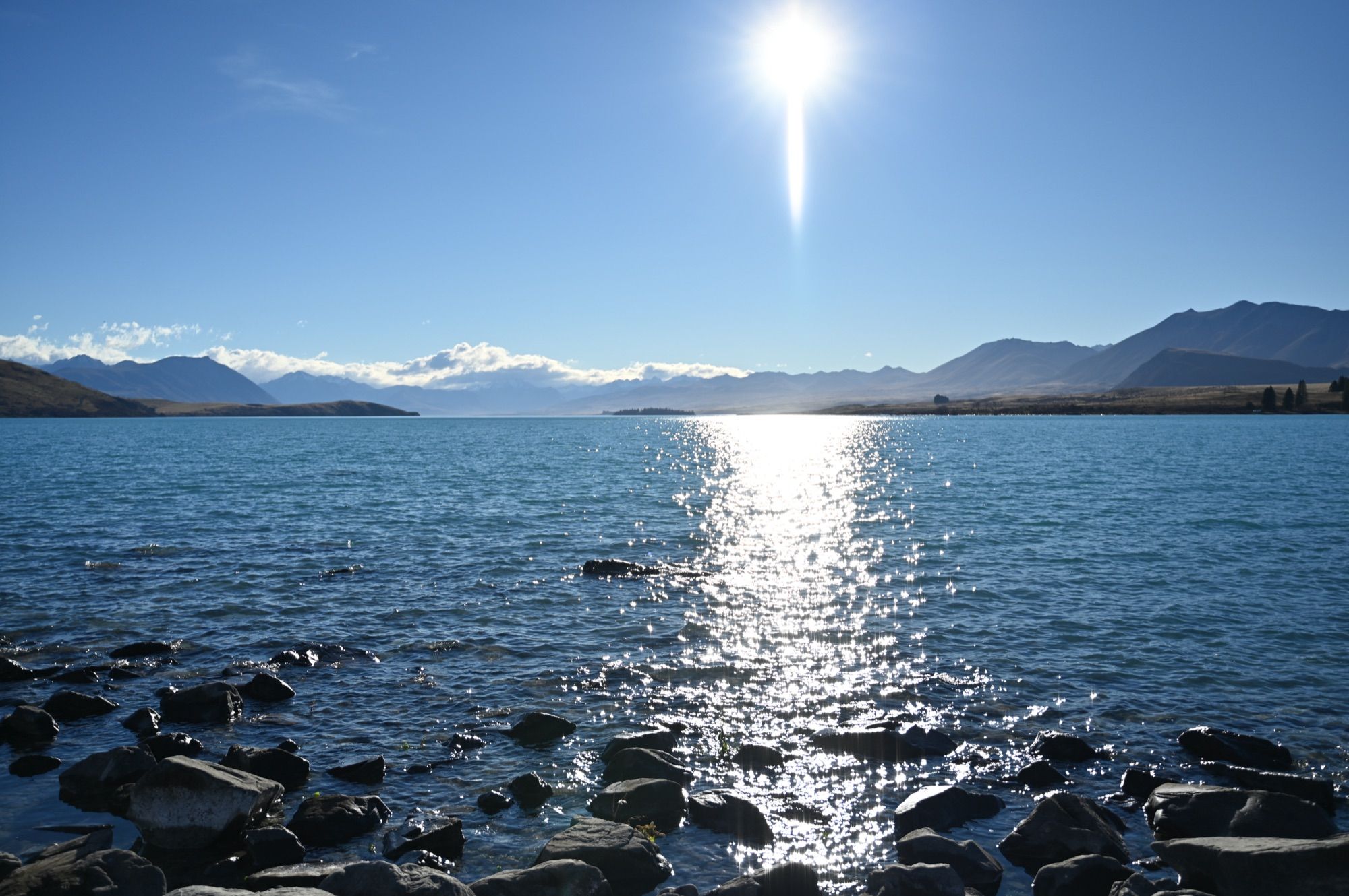 The sun blazing over Lake Tekapo, with a bright streak of light on the water and mountains on the far shore.