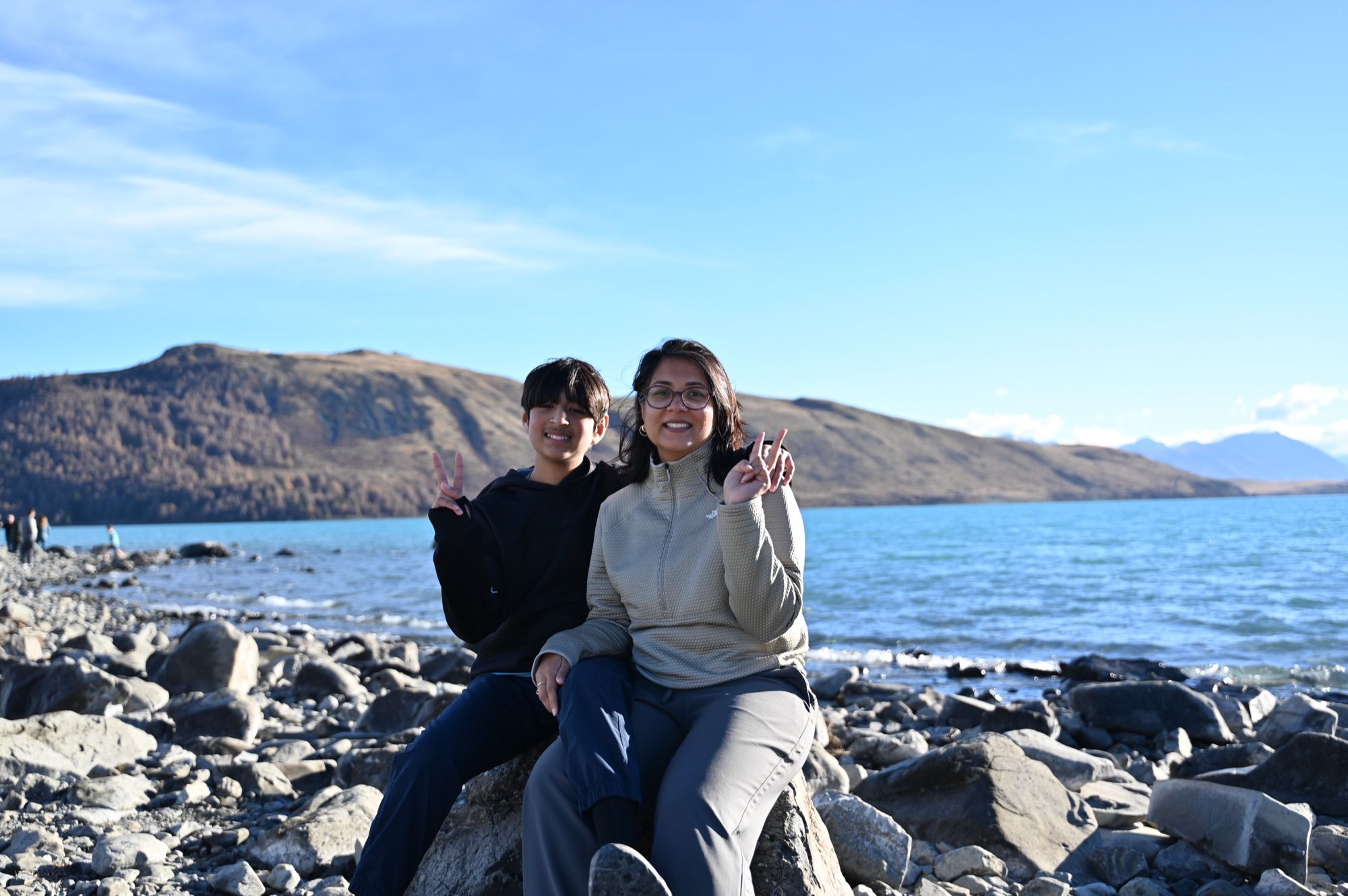Our son and I sitting on rocks at the edge of Lake Tekapo, with hills and blue water behind us.