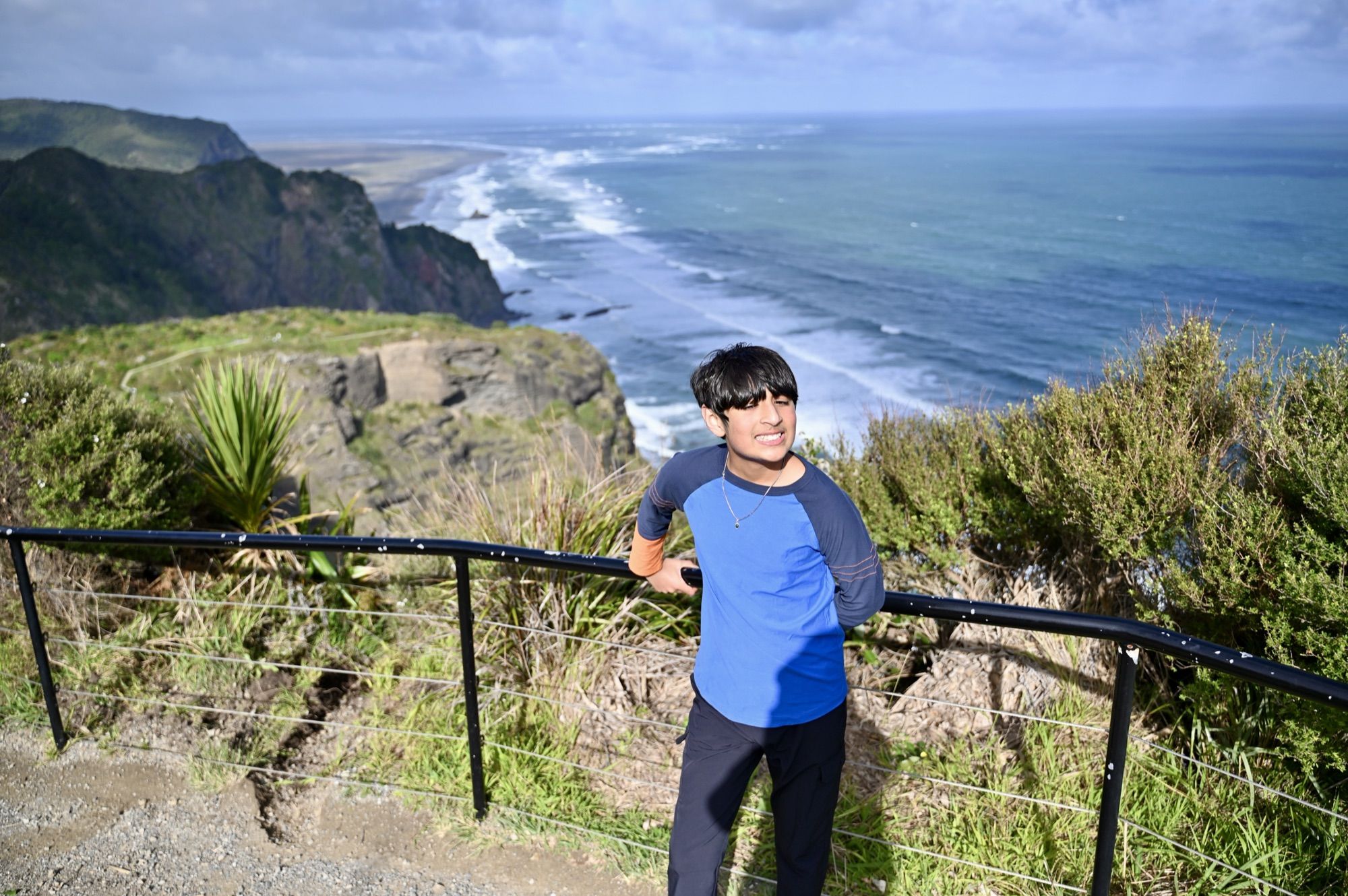 Our son leaning on the railing at a clifftop lookout, with the Tasman Sea and a long surf beach stretching behind him.