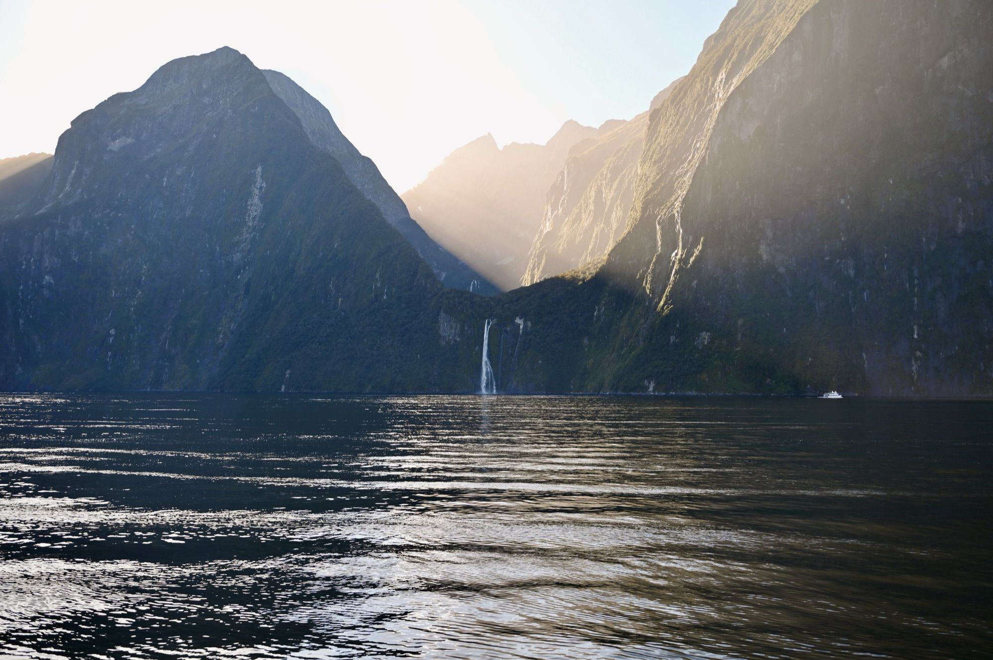 Steep, dark fiord walls rising from the water on a hazy, sunlit morning, with a thin waterfall in the distance.