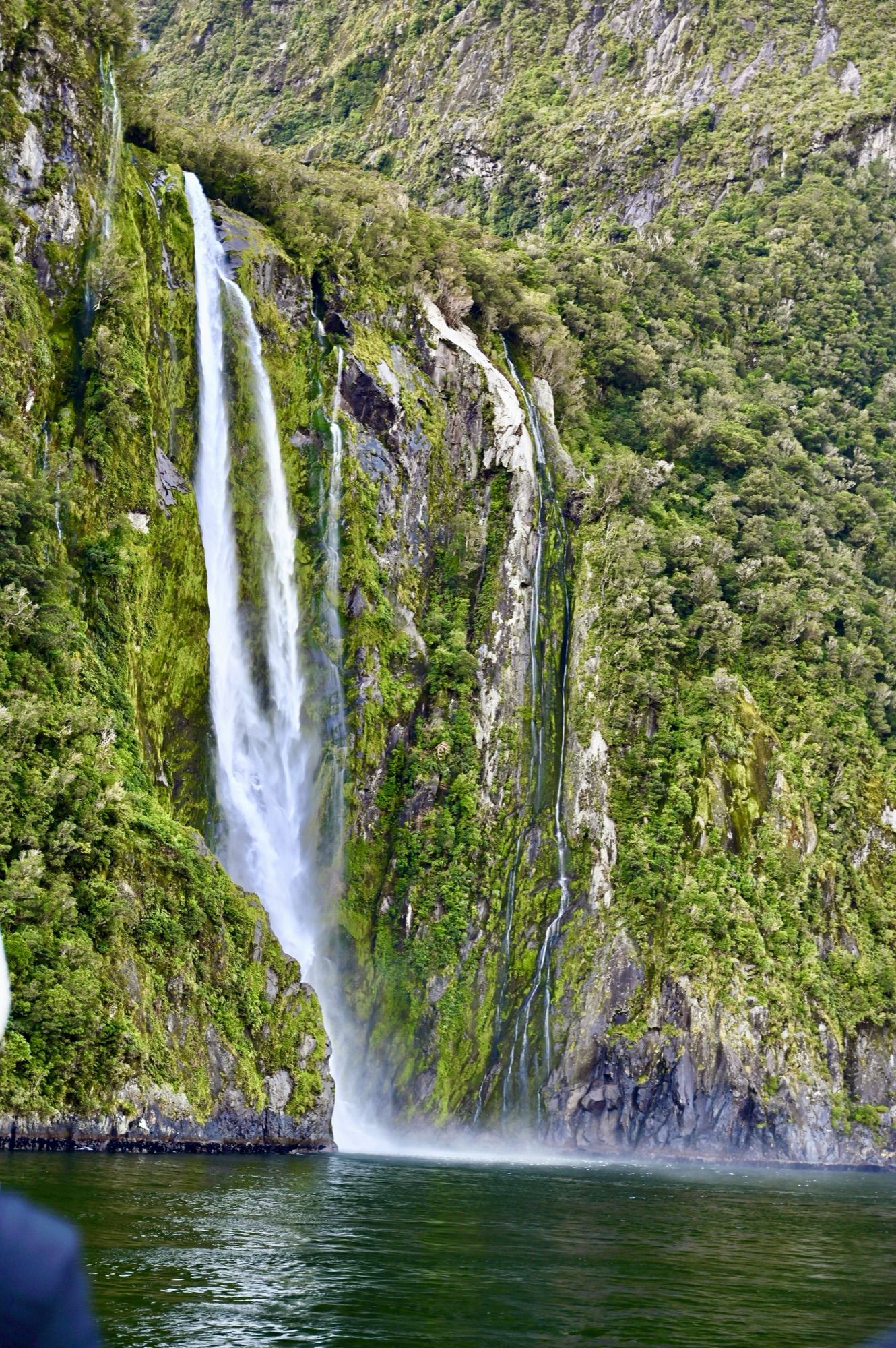 A tall waterfall plunging down a mossy green cliff straight into the fiord.