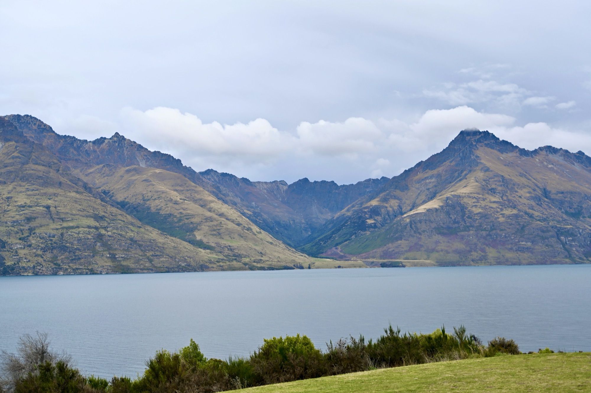 Lake Wakatipu and jagged brown mountains under a cloudy sky, with a green lawn in the foreground.