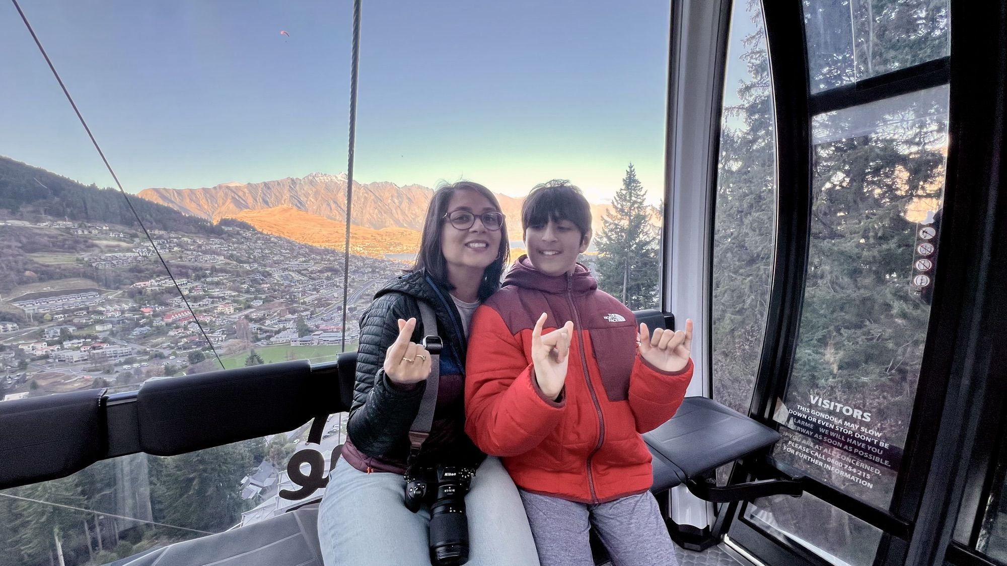 Me and our son making a hand sign inside a gondola cabin, with Queenstown and snow-dusted mountains in the window behind us.