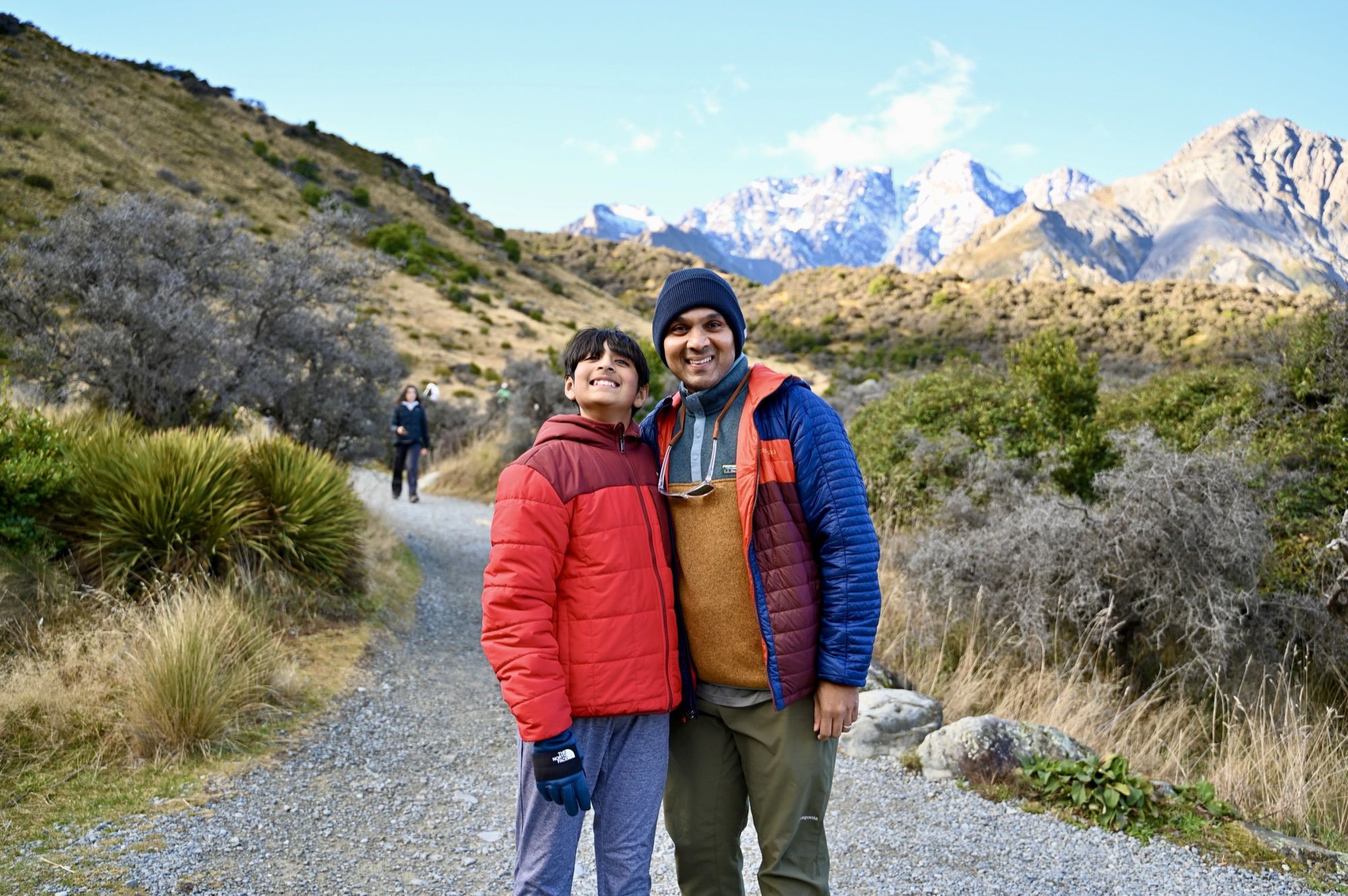 Our son and his dad smiling on a gravel path, with jagged snow-covered peaks behind them.