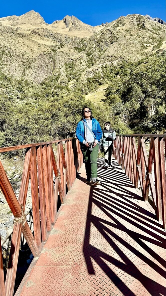 Woman walking across a metal footbridge at the start of the trail, with steep hills behind