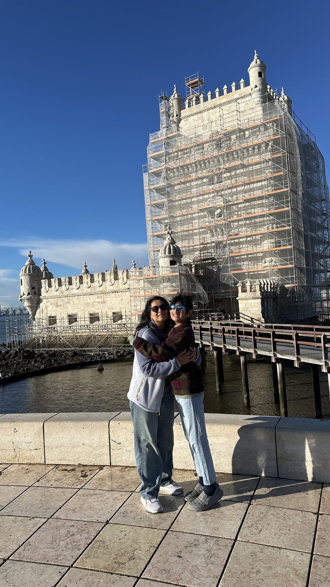 Two of us hugging on the waterfront in front of the Belém Tower, which is wrapped in scaffolding.