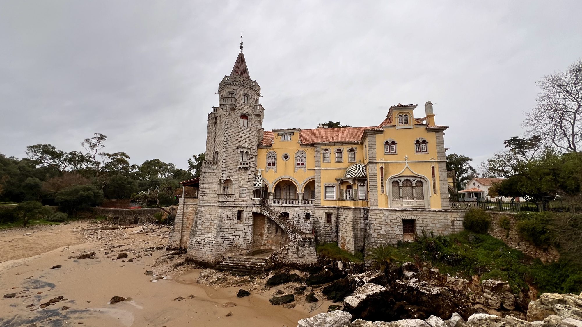 A turreted stone and yellow mansion on the rocks above a small sandy cove in Cascais.