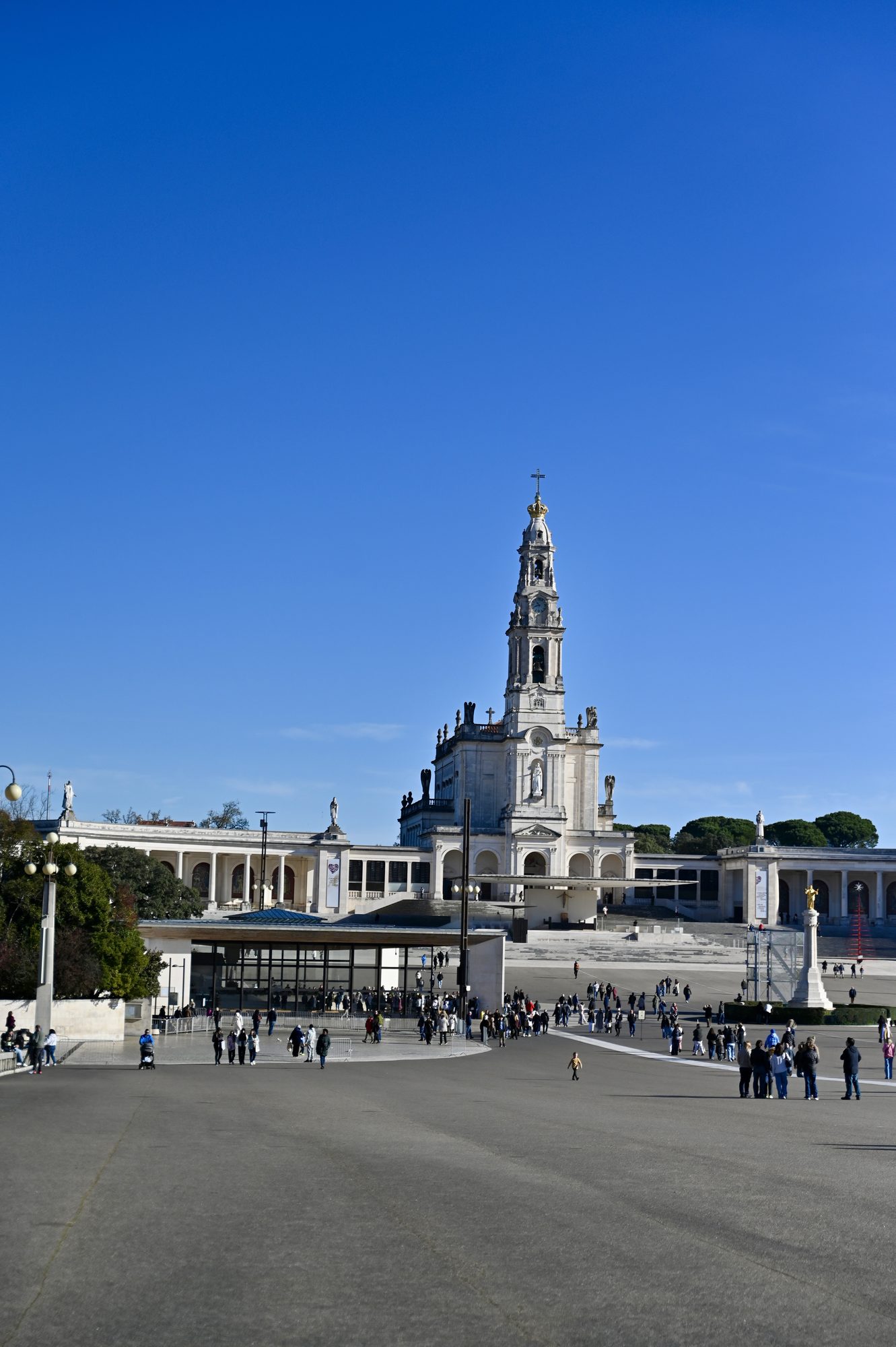 The white basilica and bell tower of Fátima at the end of a vast open square, under a deep blue sky.