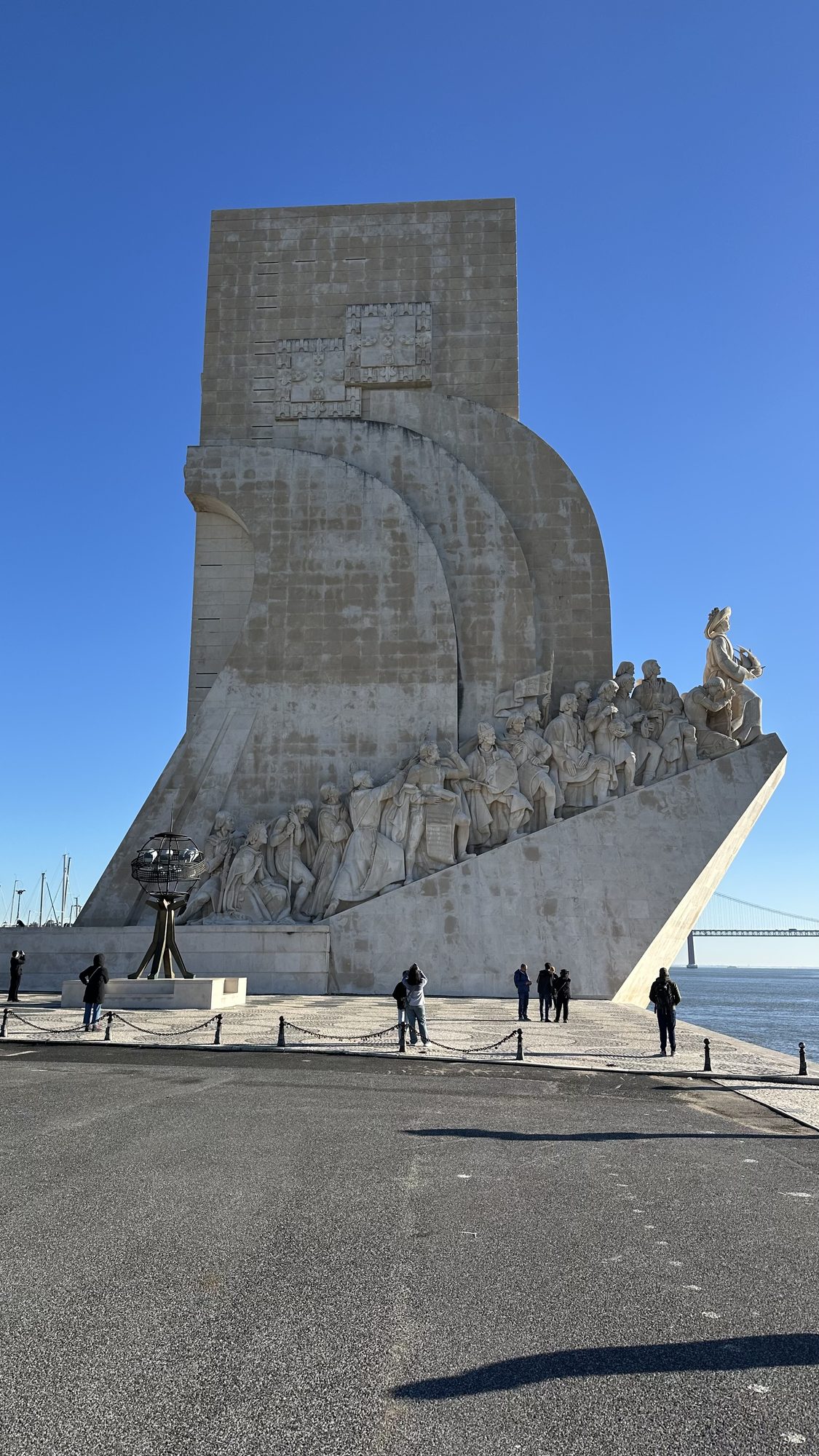 The Monument to the Discoveries, a tall white stone prow lined with carved explorers, beside the Tagus with the 25 de Abril Bridge in the distance.