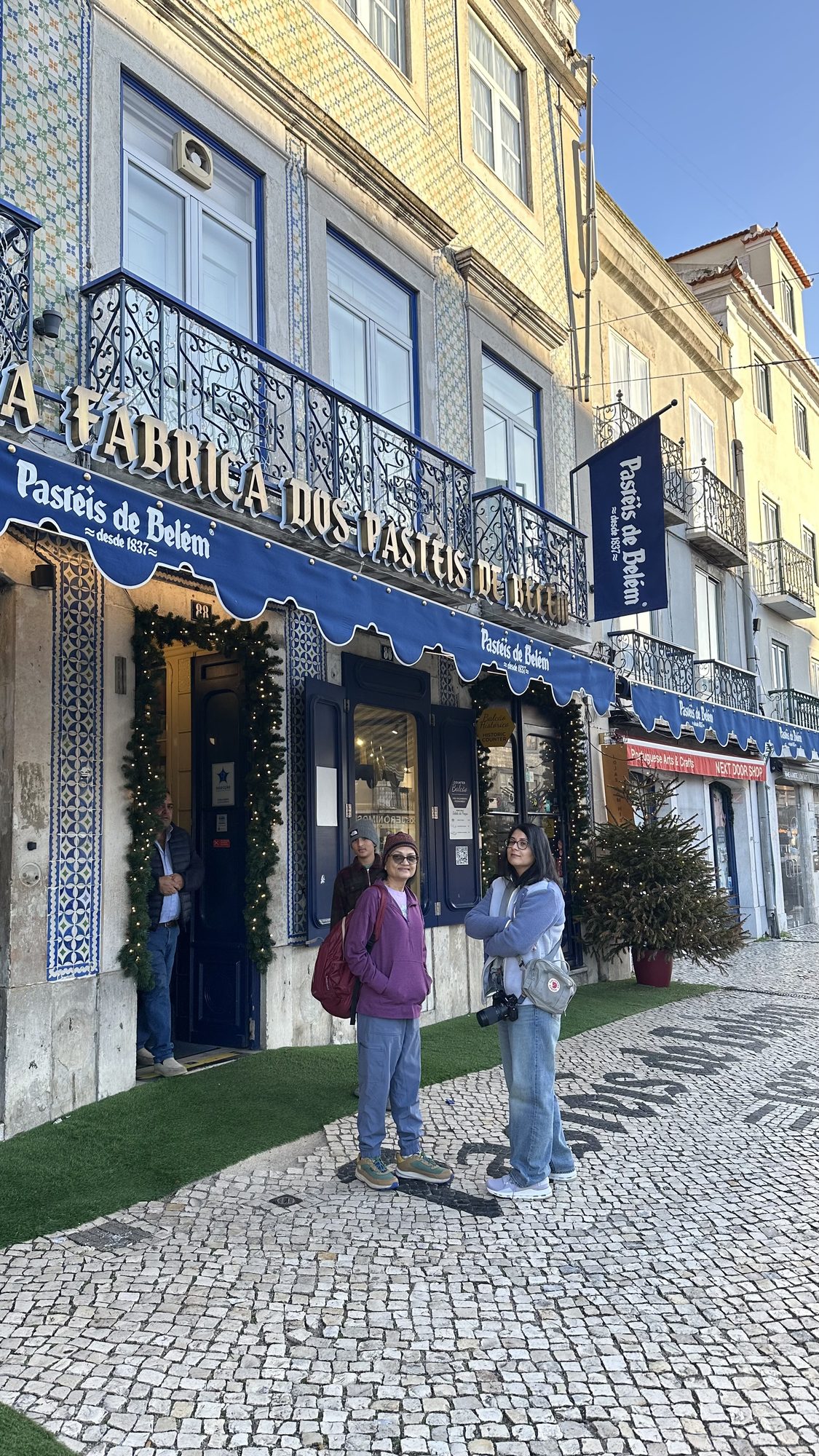 Two of us standing on the cobbled pavement outside Pastéis de Belém, under its blue awning and tiled facade with Christmas garlands.