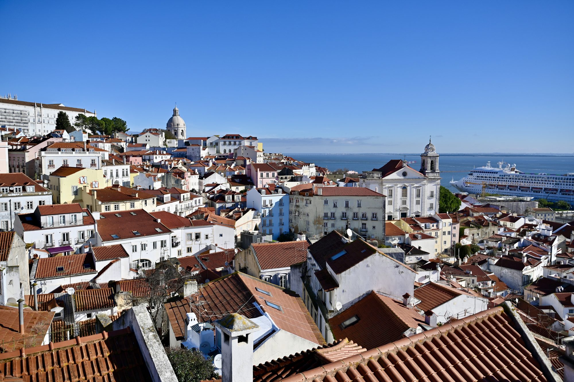 Red-tiled rooftops, church towers and a white dome stretching down to the Tagus, with a cruise ship docked in the harbor.