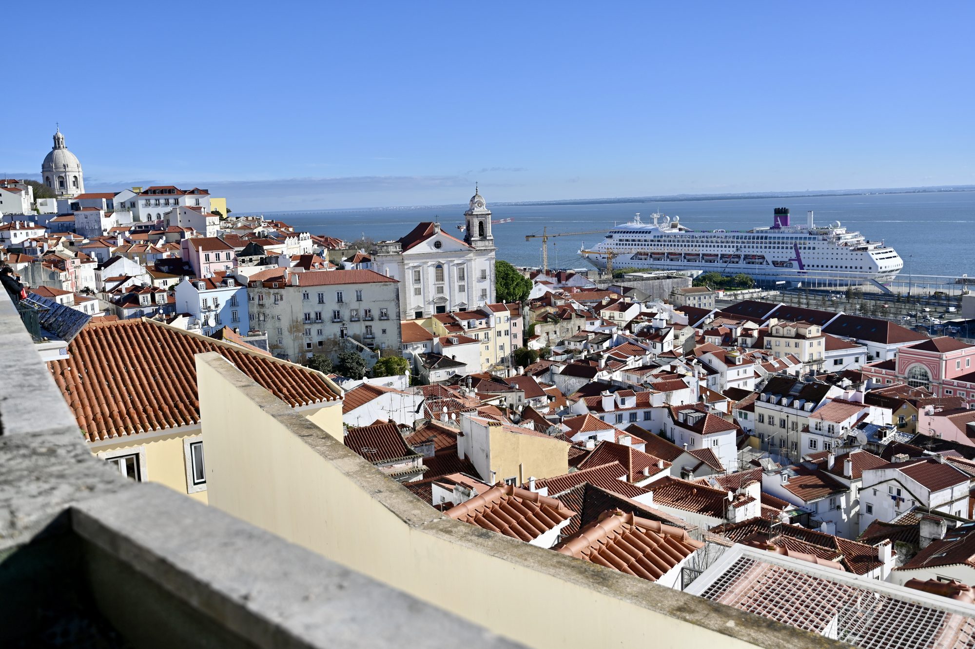 Red-tiled rooftops and church domes of Alfama stretching down to the Tagus, with a large cruise ship docked in the harbor.