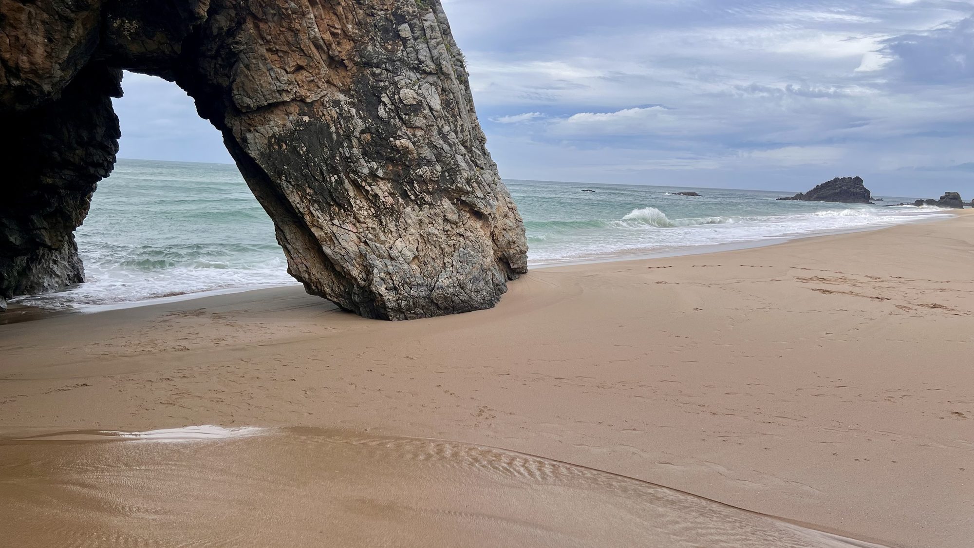 A huge rock arch standing on the sand at Praia da Adraga, with waves rolling in behind.