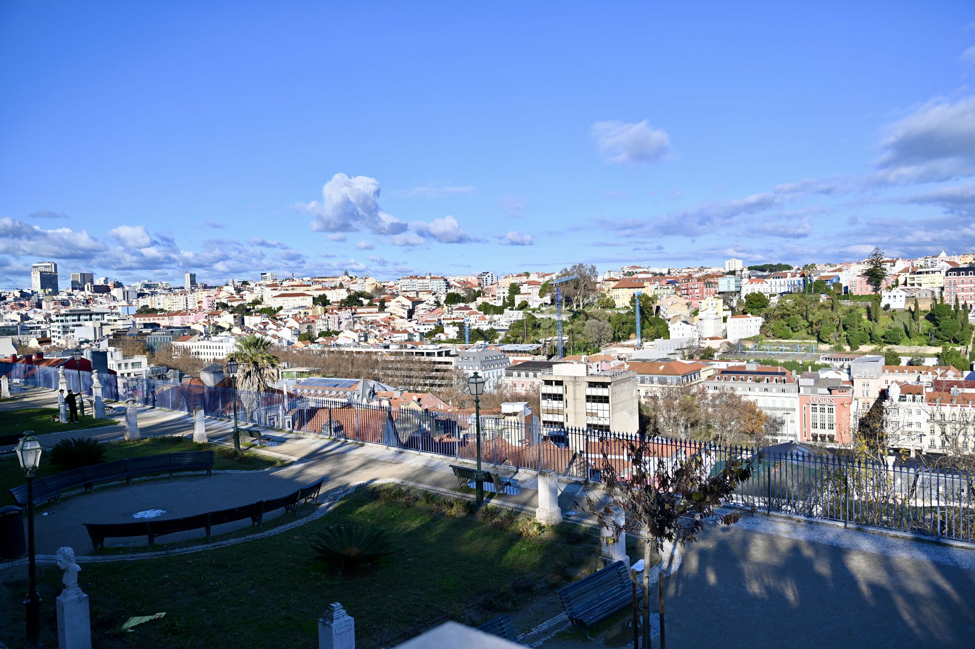 A terraced garden with paths, benches and white statues, looking out over the city under a blue sky.